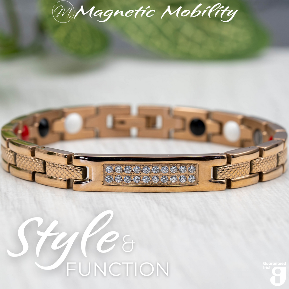 Front view of the Honesty Dawn 4in1 Magnetic Bracelet from Magnetic Mobility in rose gold finish. Features a central panel with sparkling crystals and includes Neodymium Magnets, FIR elements, Germanium, and Negative Ions for therapeutic benefits. The bracelet is shown in front of a green plant.